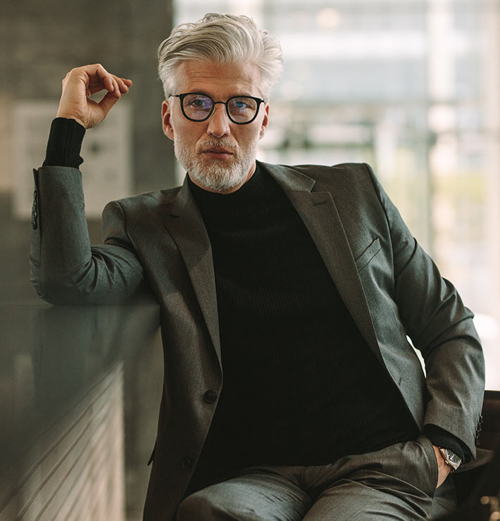 A man with glasses and his hand in pocket, sitting and posing for the photo.