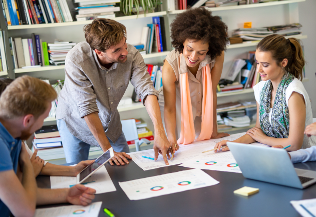 Five people in an office with books in the background, three sitting, all smiling and looking at a monitor and graphs on papers on the desk.